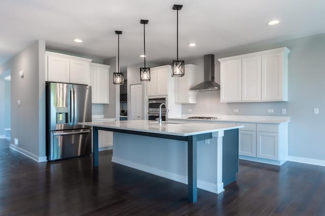 kitchen featuring a center island with sink, stainless steel appliances, light countertops, white cabinetry, and wall chimney range hood