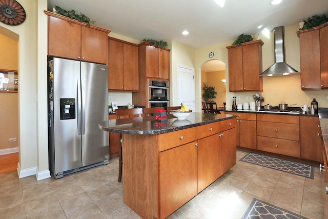 kitchen with arched walkways, a center island, stainless steel appliances, brown cabinetry, and wall chimney exhaust hood