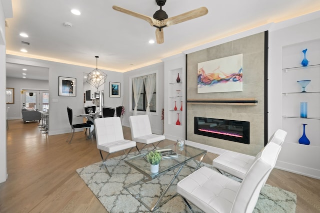 living area featuring built in shelves, a tile fireplace, ceiling fan with notable chandelier, baseboards, and light wood-style floors