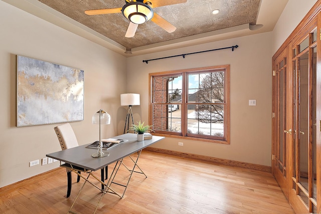 home office featuring light wood-style floors, a tray ceiling, baseboards, and a ceiling fan