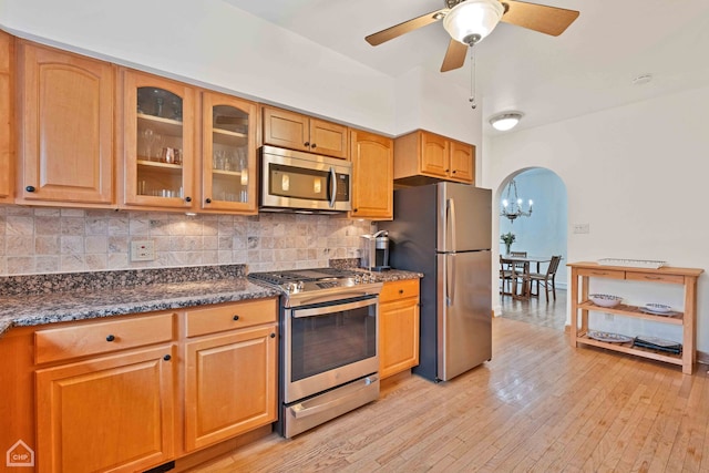 kitchen featuring arched walkways, stainless steel appliances, tasteful backsplash, light wood-style floors, and glass insert cabinets