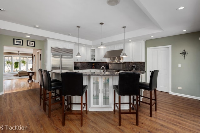 kitchen with stainless steel built in fridge, dark wood-type flooring, tasteful backsplash, range hood, and glass insert cabinets