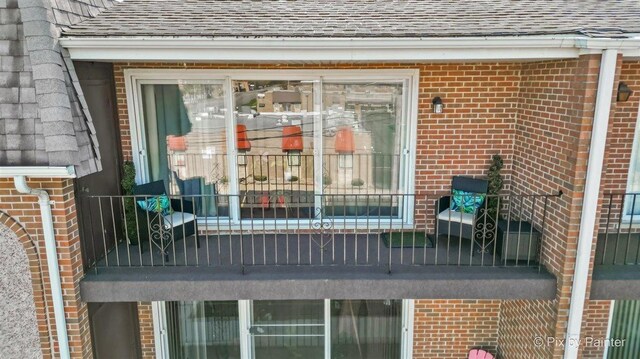 entrance to property featuring a shingled roof, a balcony, and brick siding