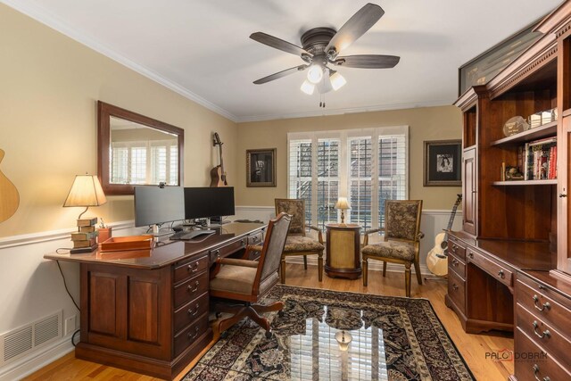 home office with a ceiling fan, a wainscoted wall, visible vents, light wood finished floors, and ornamental molding