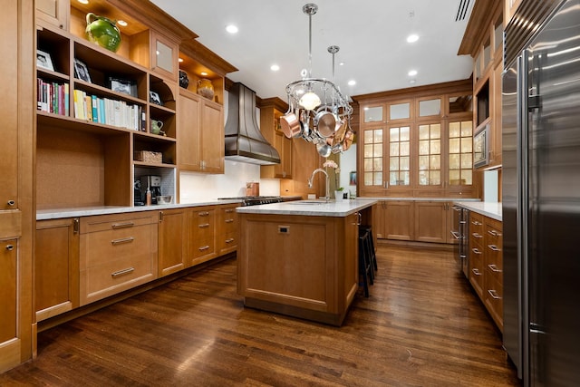 kitchen with custom exhaust hood, open shelves, a kitchen island with sink, a sink, and built in appliances