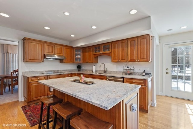 kitchen with black electric stovetop, under cabinet range hood, stainless steel dishwasher, brown cabinets, and a kitchen bar