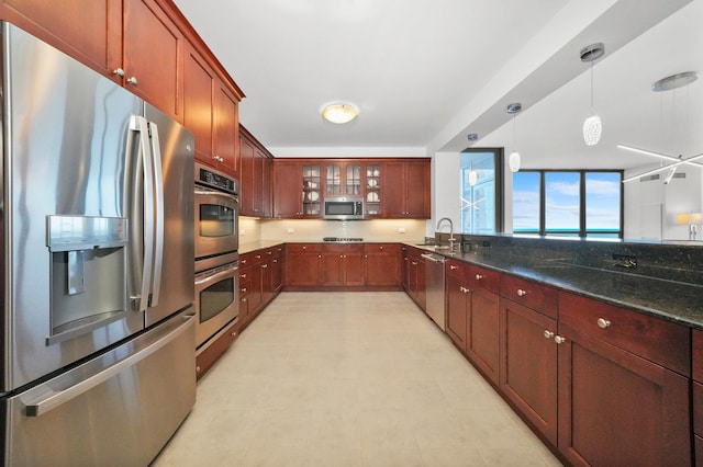 kitchen featuring pendant lighting, stainless steel appliances, glass insert cabinets, a sink, and dark stone counters