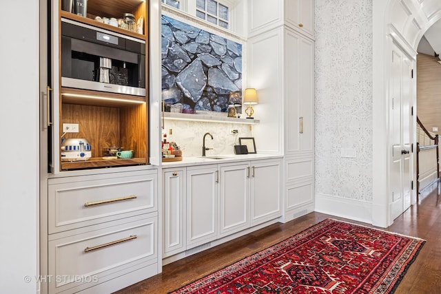 bar featuring wallpapered walls, wet bar, arched walkways, a sink, and dark wood-type flooring