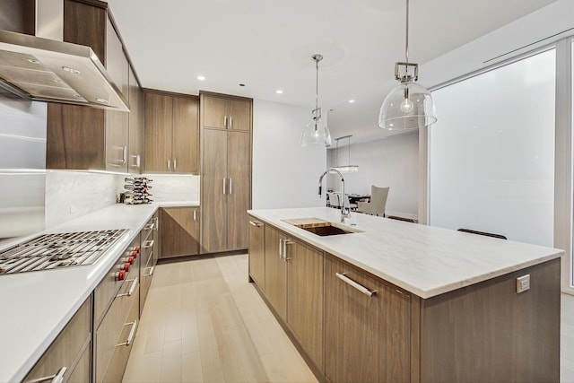 kitchen featuring light wood-style flooring, an island with sink, a sink, wall chimney range hood, and stainless steel gas cooktop