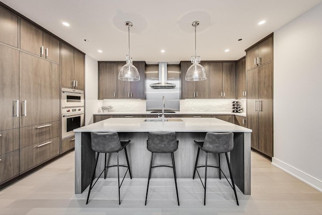 kitchen with a breakfast bar, light countertops, wall chimney range hood, and a kitchen island with sink