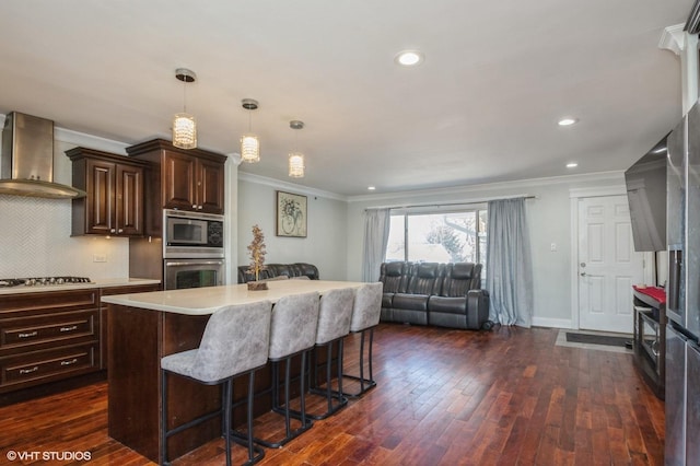 kitchen featuring wall chimney exhaust hood, appliances with stainless steel finishes, open floor plan, a breakfast bar area, and light countertops