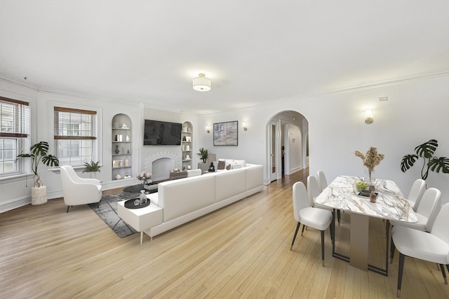 living room with arched walkways, light wood-style flooring, a fireplace, built in features, and ornamental molding