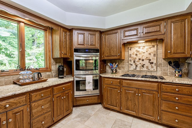 kitchen with brown cabinets, tasteful backsplash, gas stovetop, double oven, and light stone countertops