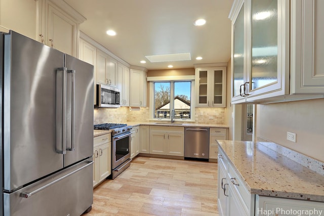 kitchen featuring high quality appliances, a sink, light stone counters, tasteful backsplash, and glass insert cabinets