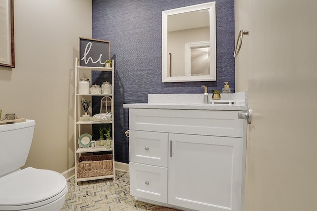 half bathroom with baseboards, vanity, toilet, and wallpapered walls