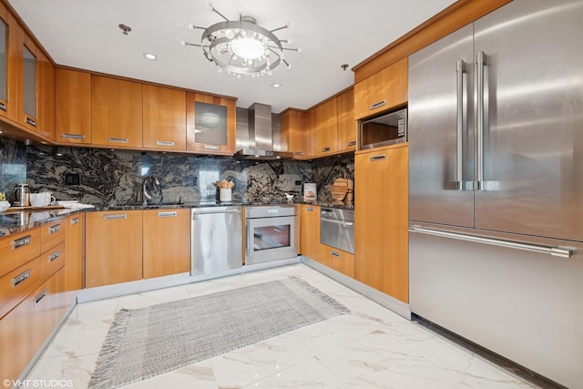 kitchen featuring marble finish floor, wall chimney exhaust hood, dark stone counters, and built in appliances