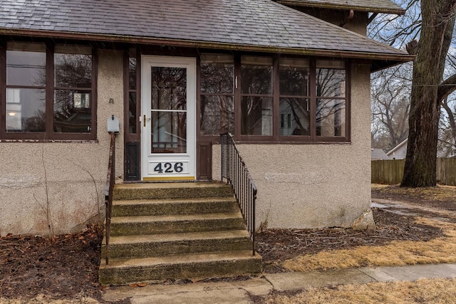 entrance to property featuring stucco siding, fence, and roof with shingles