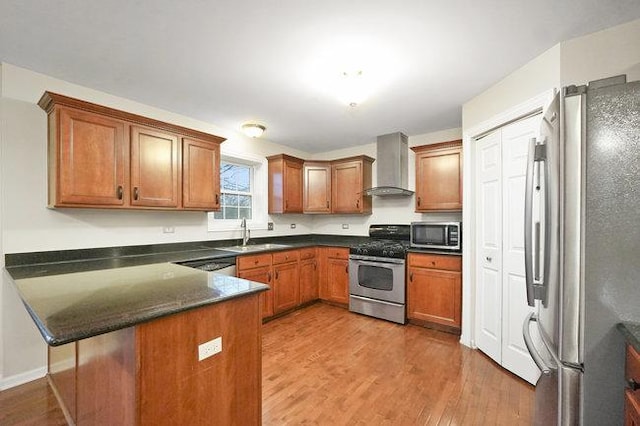 kitchen featuring wall chimney exhaust hood, brown cabinets, a peninsula, stainless steel appliances, and a sink