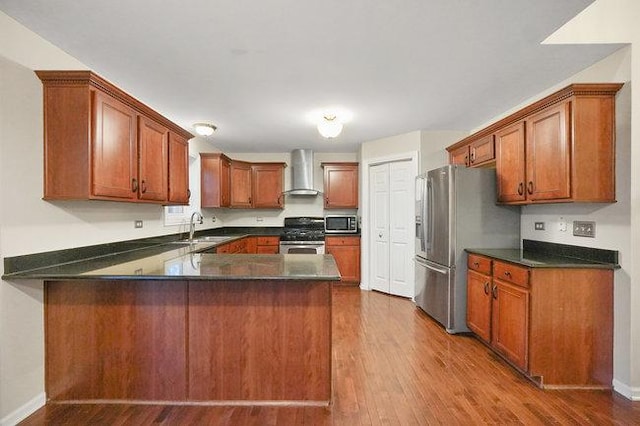 kitchen featuring dark wood finished floors, stainless steel appliances, dark countertops, wall chimney range hood, and a peninsula