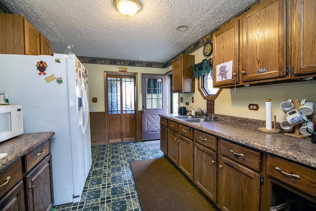 kitchen featuring white appliances, wainscoting, dark countertops, a textured ceiling, and a sink