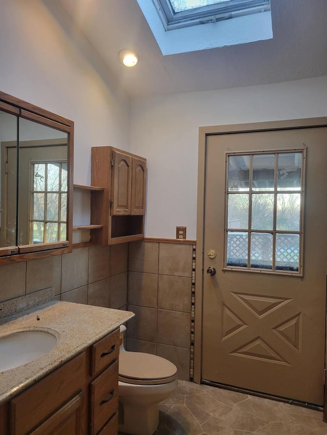 bathroom featuring toilet, a wainscoted wall, a skylight, vanity, and tile walls