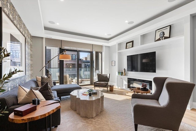 living room with a glass covered fireplace, a raised ceiling, light wood-style flooring, and baseboards