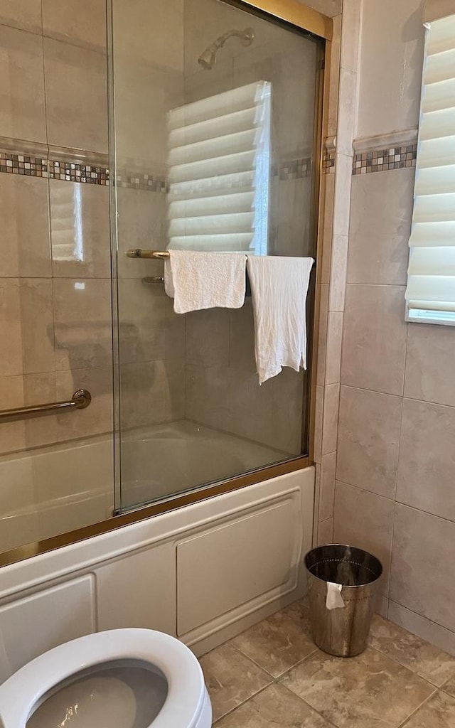 bathroom featuring toilet, combined bath / shower with glass door, and tile walls