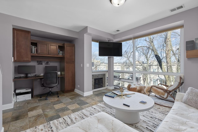 living room with a fireplace, visible vents, baseboards, built in study area, and stone tile flooring