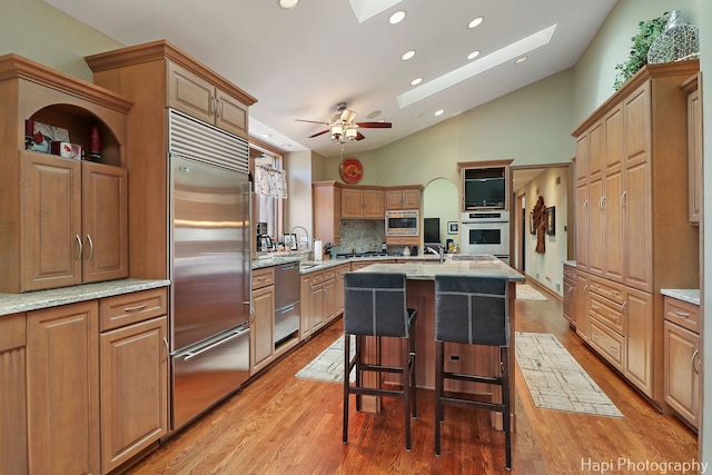 kitchen featuring light stone counters, a skylight, a kitchen island with sink, built in appliances, and light wood-type flooring