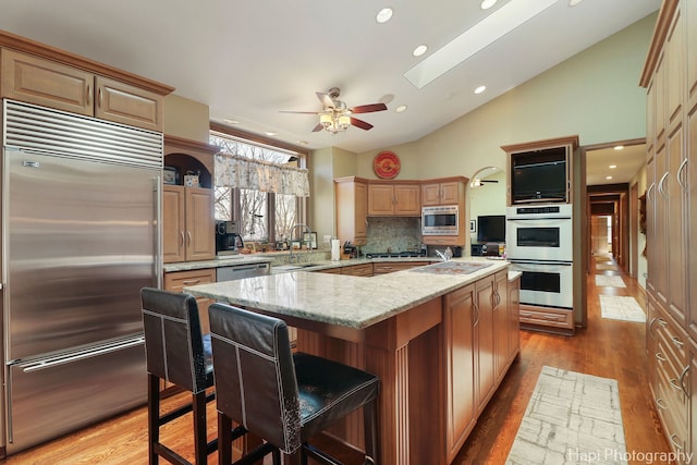 kitchen featuring light stone counters, light wood-style flooring, an island with sink, a sink, and built in appliances