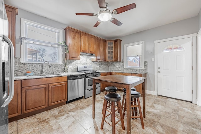 kitchen featuring tasteful backsplash, appliances with stainless steel finishes, brown cabinetry, a sink, and under cabinet range hood