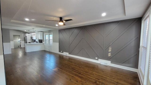 unfurnished living room with a decorative wall, a ceiling fan, visible vents, dark wood-style floors, and a raised ceiling