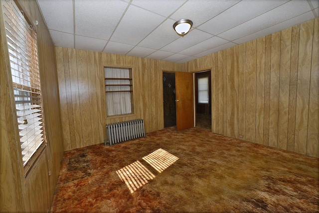 carpeted spare room with radiator, wooden walls, and a drop ceiling