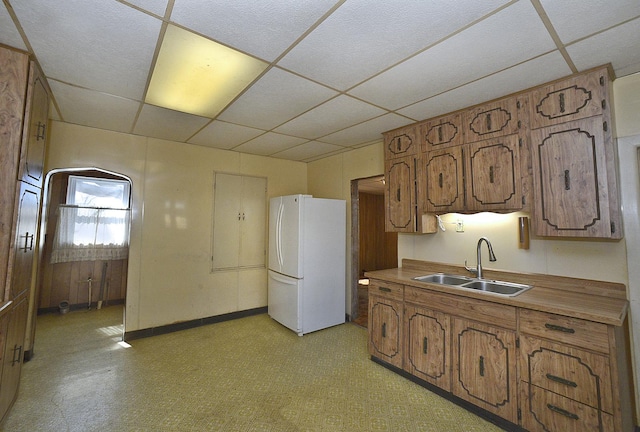 kitchen featuring brown cabinets, freestanding refrigerator, a sink, a drop ceiling, and baseboards