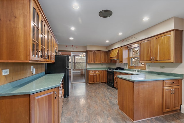 kitchen with brown cabinetry, a peninsula, black appliances, premium range hood, and a sink