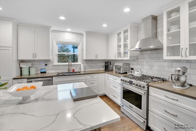 kitchen with stainless steel appliances, a sink, white cabinets, light wood-type flooring, and wall chimney exhaust hood