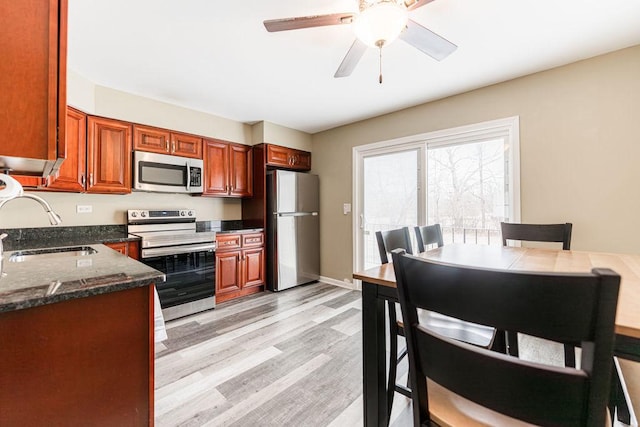 kitchen featuring stainless steel appliances, a sink, a ceiling fan, light wood-type flooring, and dark stone countertops