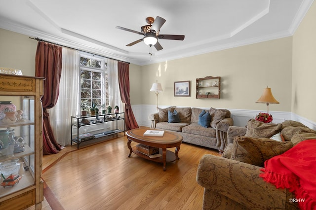 living area with a ceiling fan, wood finished floors, ornamental molding, wainscoting, and a raised ceiling