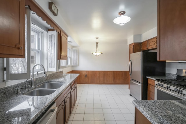 kitchen with pendant lighting, a wainscoted wall, stainless steel appliances, brown cabinetry, and a sink