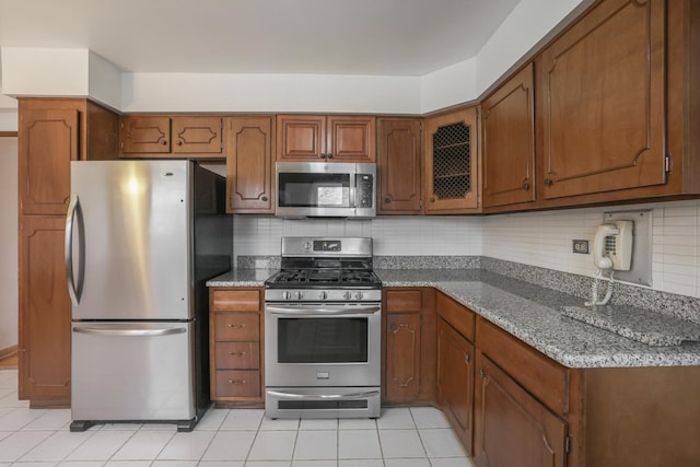 kitchen featuring light tile patterned floors, tasteful backsplash, appliances with stainless steel finishes, brown cabinets, and dark stone countertops