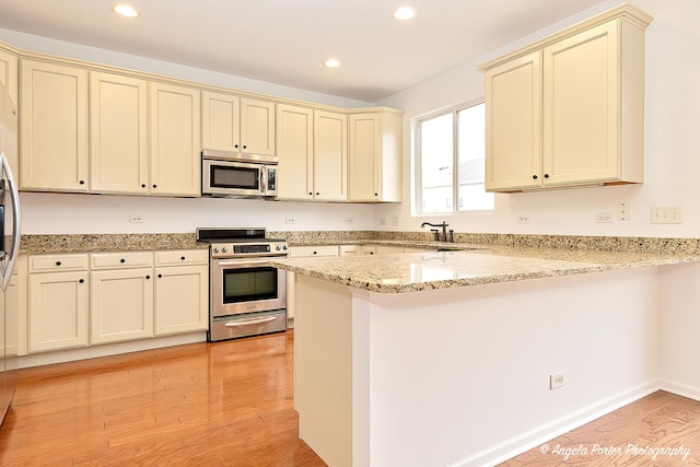 kitchen with light stone counters, cream cabinetry, appliances with stainless steel finishes, light wood-type flooring, and a peninsula