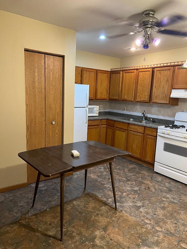 kitchen featuring dark countertops, brown cabinetry, a sink, white appliances, and under cabinet range hood