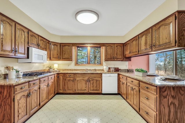 kitchen with white appliances, a sink, light stone countertops, light floors, and brown cabinetry