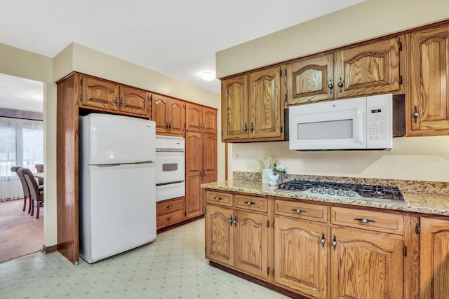 kitchen featuring light stone counters, a warming drawer, light floors, brown cabinetry, and white appliances