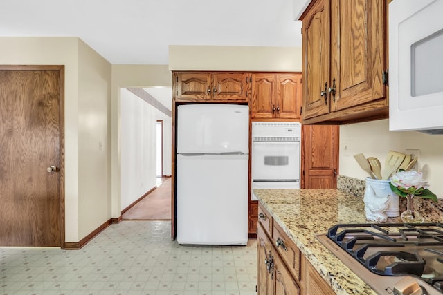 kitchen featuring white appliances, baseboards, brown cabinets, light stone countertops, and light floors