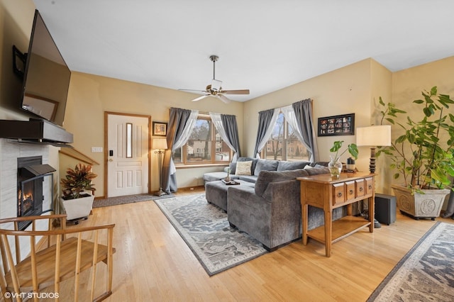 living area with light wood-type flooring, a warm lit fireplace, and a ceiling fan