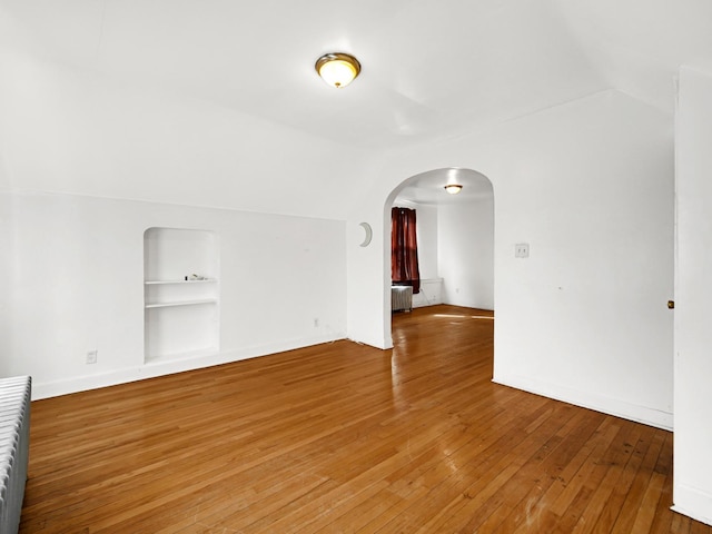 unfurnished room featuring built in features, baseboards, arched walkways, vaulted ceiling, and wood-type flooring