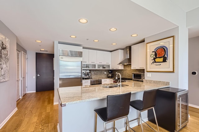 kitchen featuring tasteful backsplash, a peninsula, stainless steel built in refrigerator, wall chimney range hood, and a sink