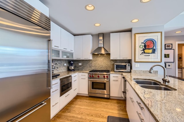 kitchen with built in appliances, a sink, backsplash, wall chimney exhaust hood, and light wood finished floors