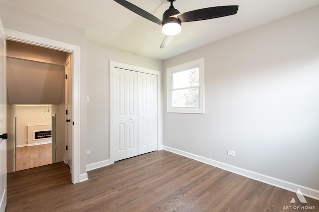 unfurnished bedroom featuring a warm lit fireplace, baseboards, a ceiling fan, wood finished floors, and a closet